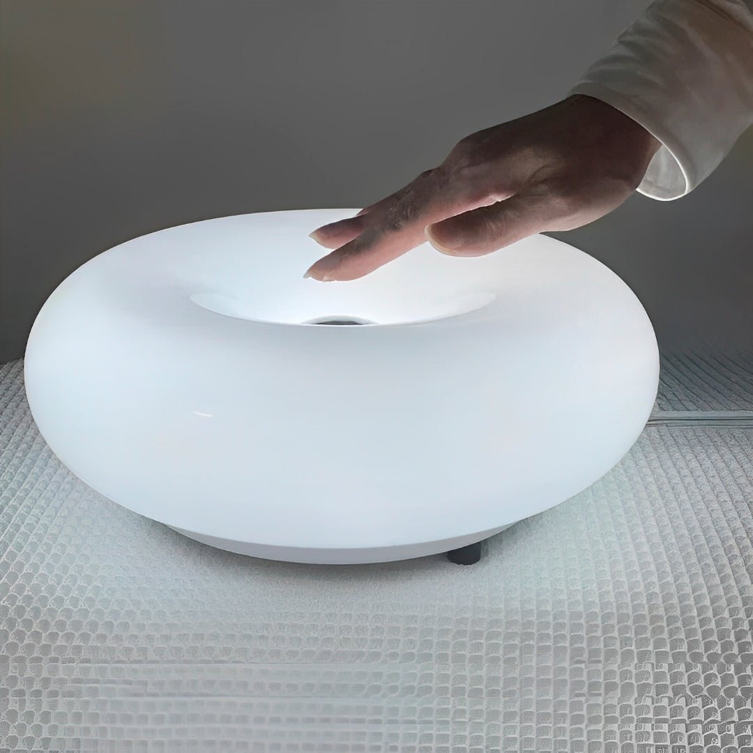 Mão tocando o alto da luminária donut branca acesa, sobre toalha clara