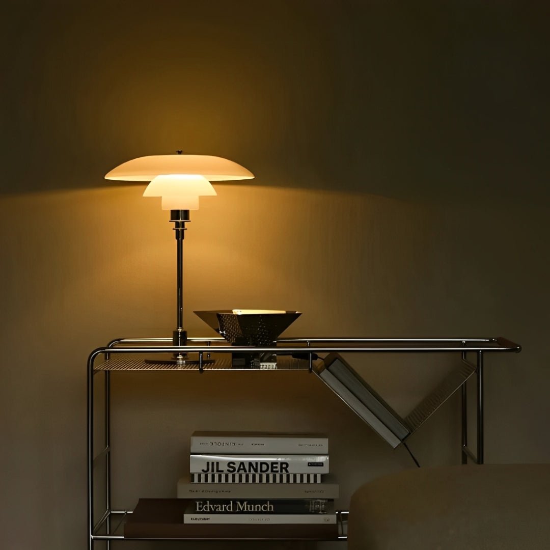 Abajur aceso sobre mesa de metal e vidro, com tigela ao lado e livros embaixo