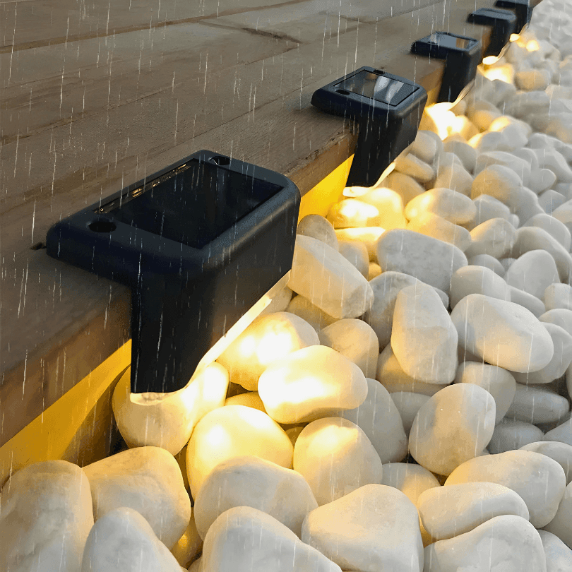 Luminárias solares acesas na borda de um deck, sobre pedras brancas, sob chuva fina