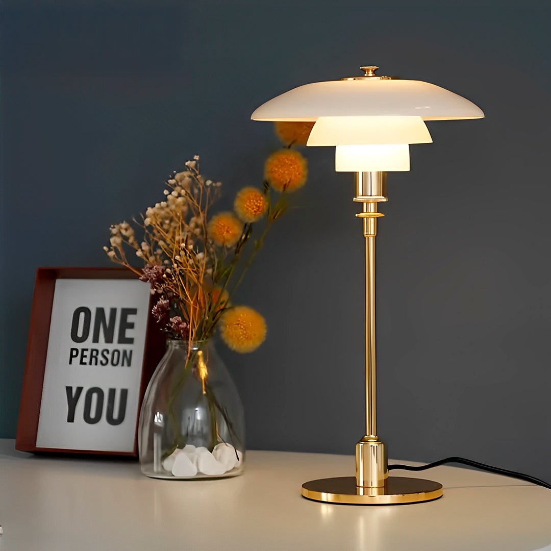 Abajur sem fio dourado aceso sobre mesa branca, ao lado de vaso com flores secas