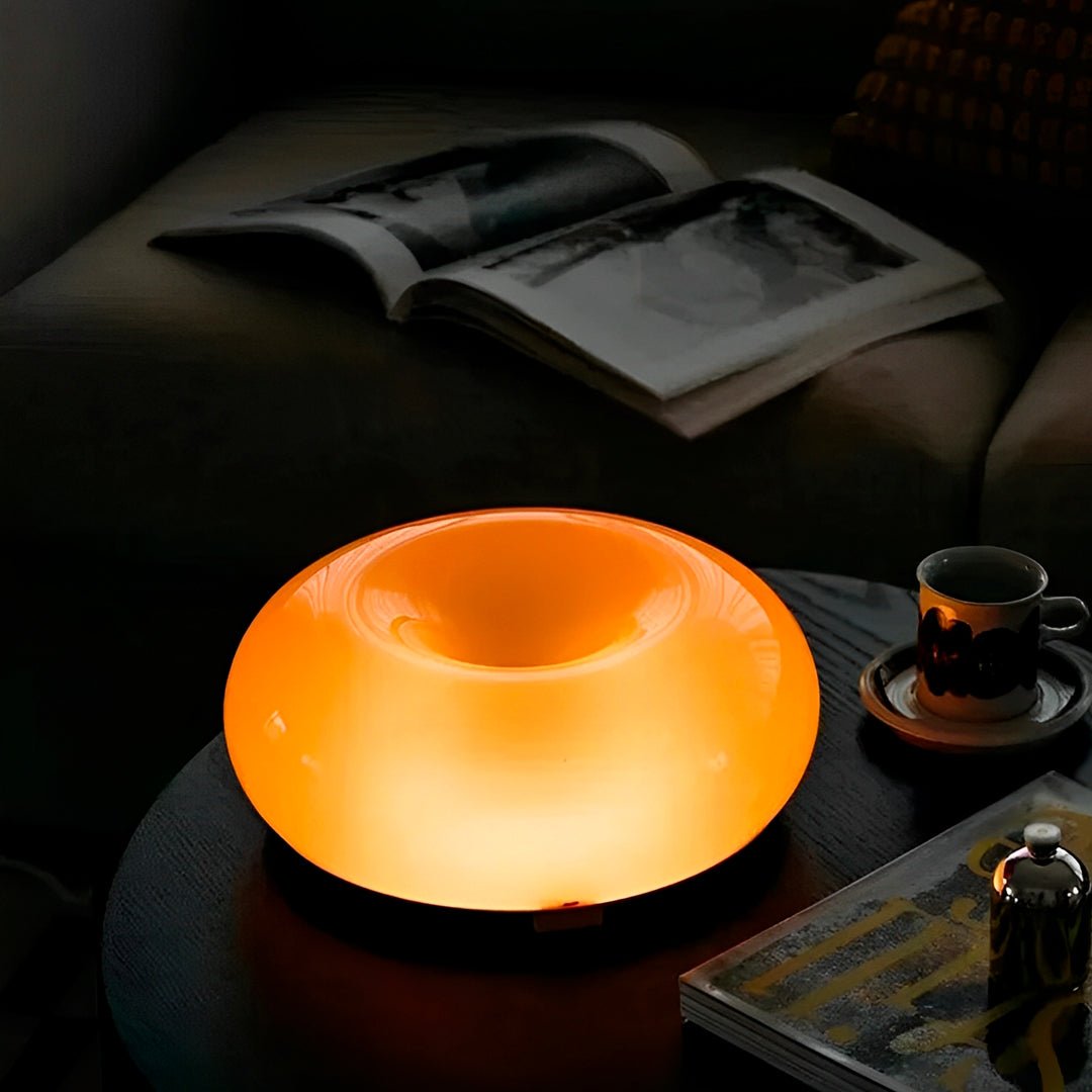 Luminária donut laranja acesa em mesa lateral escura, ao lado de xícara e revista