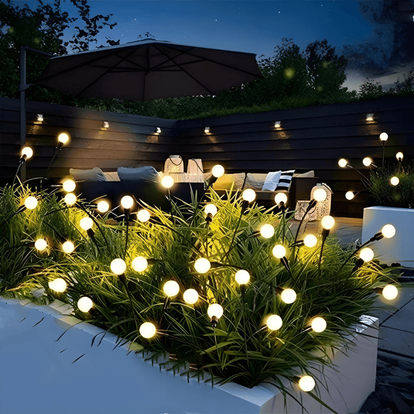 Luminária vagalume solar acesa entre plantas de um canteiro, com área de estar ao fundo