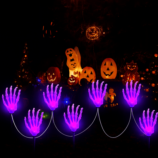 Mãos de esqueleto acesas em roxo no jardim, ao lado de abóboras e fantasmas decorativos
