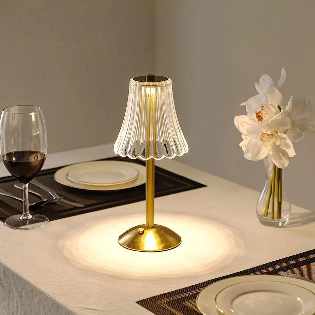 Abajur dourado portátil aceso sobre mesa de jantar posta, com taça, pratos e flores brancas