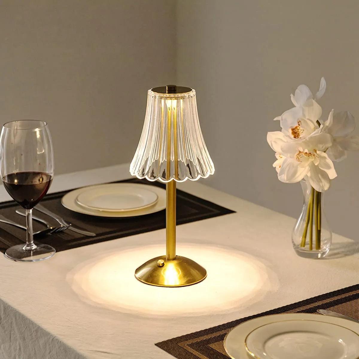Abajur dourado portátil aceso sobre mesa de jantar posta, com taça, pratos e flores brancas