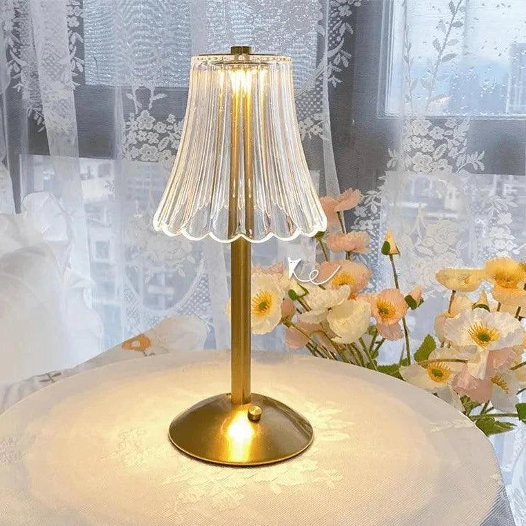 Abajur dourado aceso sobre mesa redonda com toalha, flores e cortina de renda ao fundo