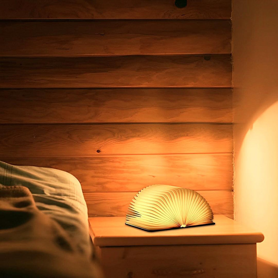 Luminária de livro aberta em meia-lua e acesa sobre o criado-mudo, ao lado da cama
