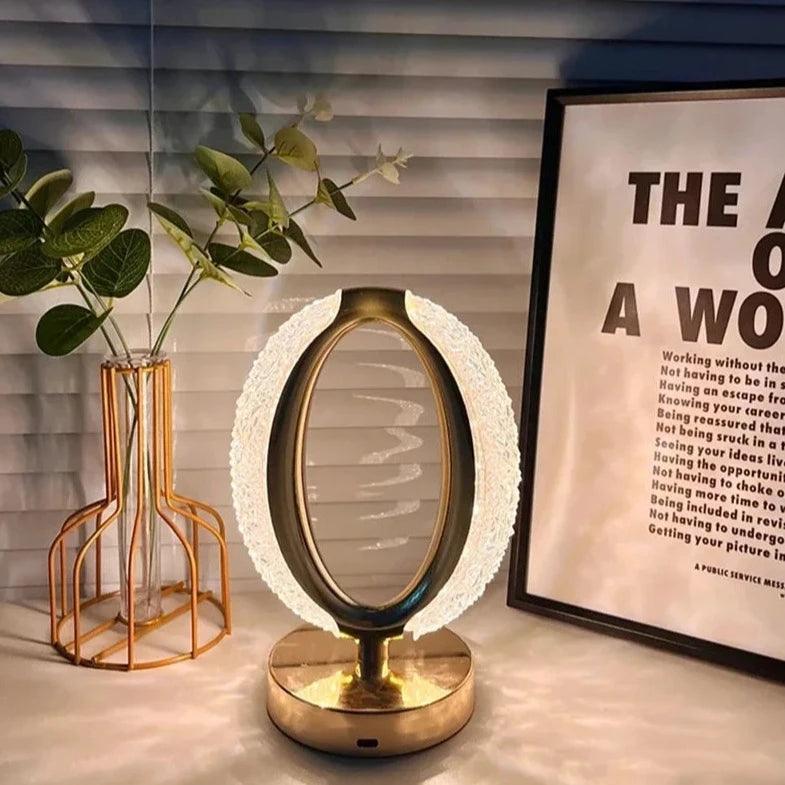 Luminária de mesa dourada acesa, com anel de acrílico, ao lado de vaso e quadro