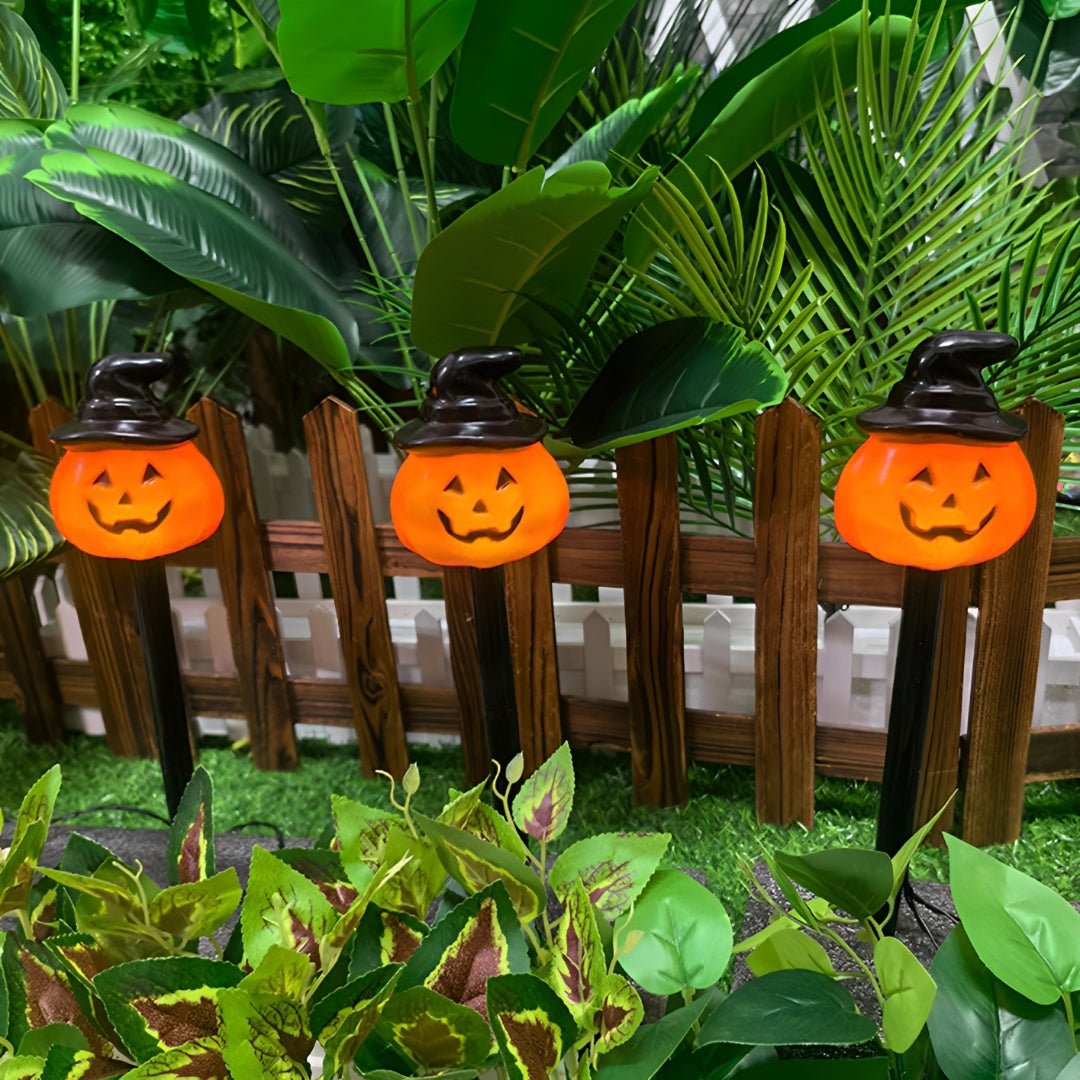 Três abóboras com chapéu de bruxa acesas, fincadas entre plantas junto a uma cerca