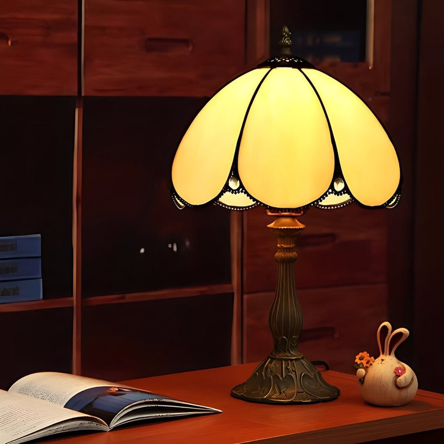 Abajur Tiffany aceso sobre mesa de madeira, com cúpula de vitral amarelada e livro aberto