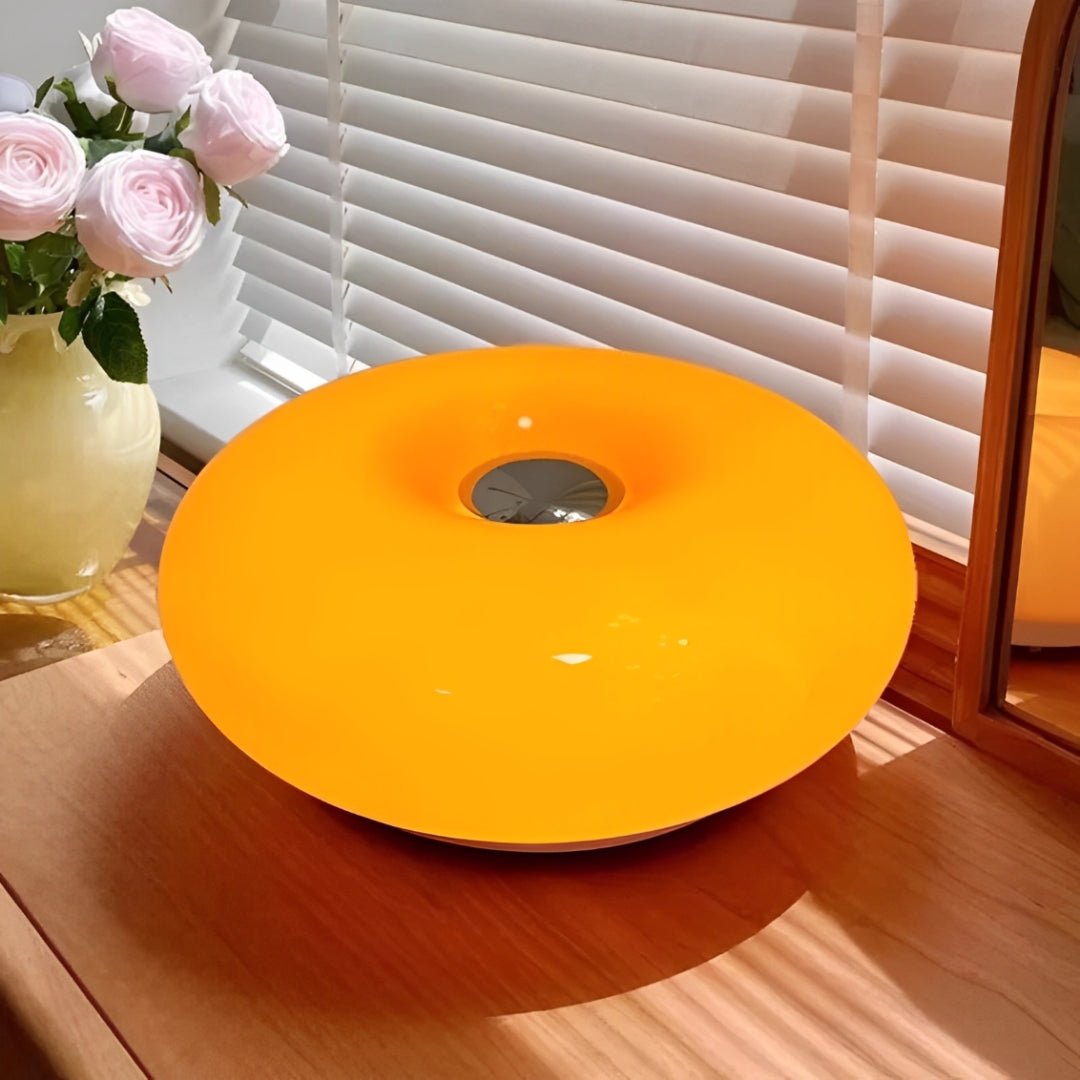 Luminária donut de vidro laranja vista de cima sobre móvel de madeira, com flores