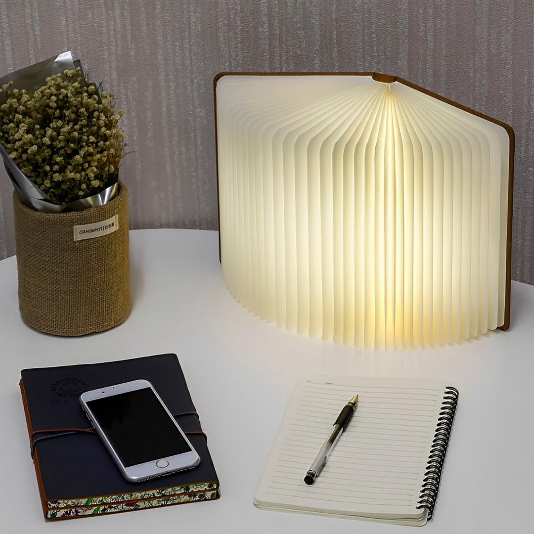 Luminária livro em pé e aberta sobre a mesa, ao lado de caderno e celular