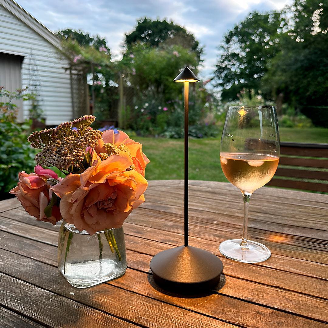 Luminária de mesa sem fio estilo Pina acesa numa mesa de jardim, junto de flores
