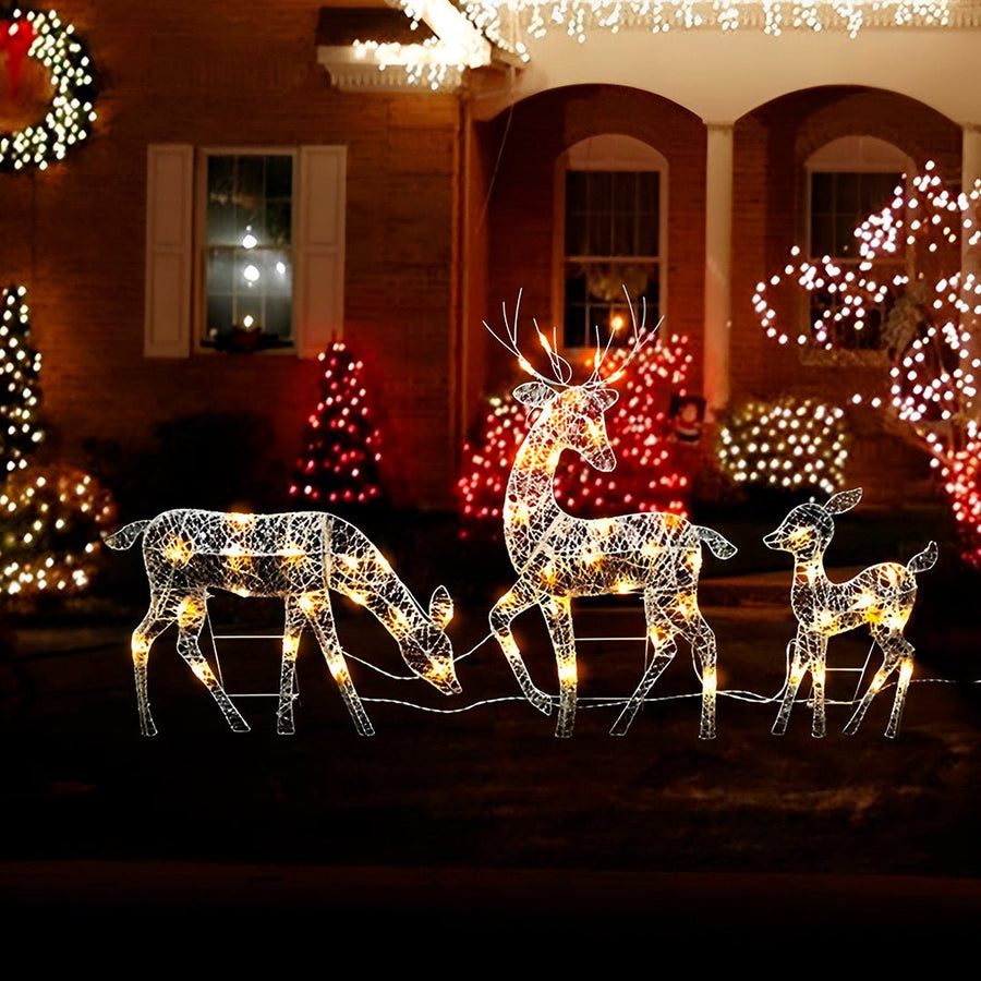 Família de renas acesas diante de uma casa decorada com luzes de Natal, à noite