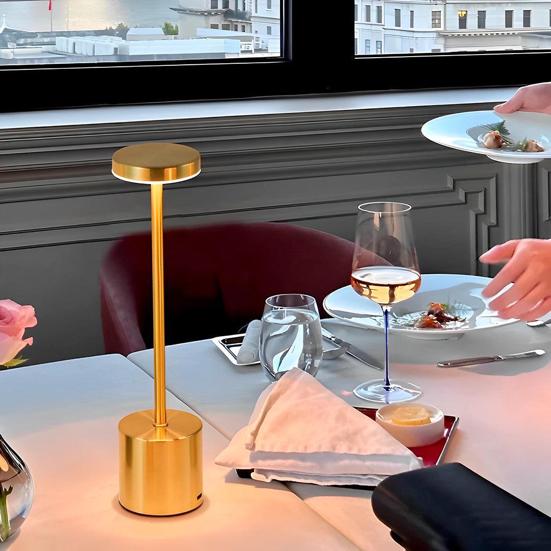 Luminária dourada acesa em mesa de restaurante, ao lado de taça, prato e guardanapo