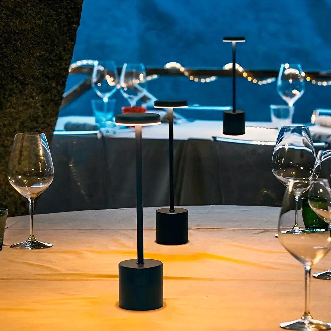 Luminária preta acesa em mesa à beira-mar, entre taças de vinho, ao anoitecer