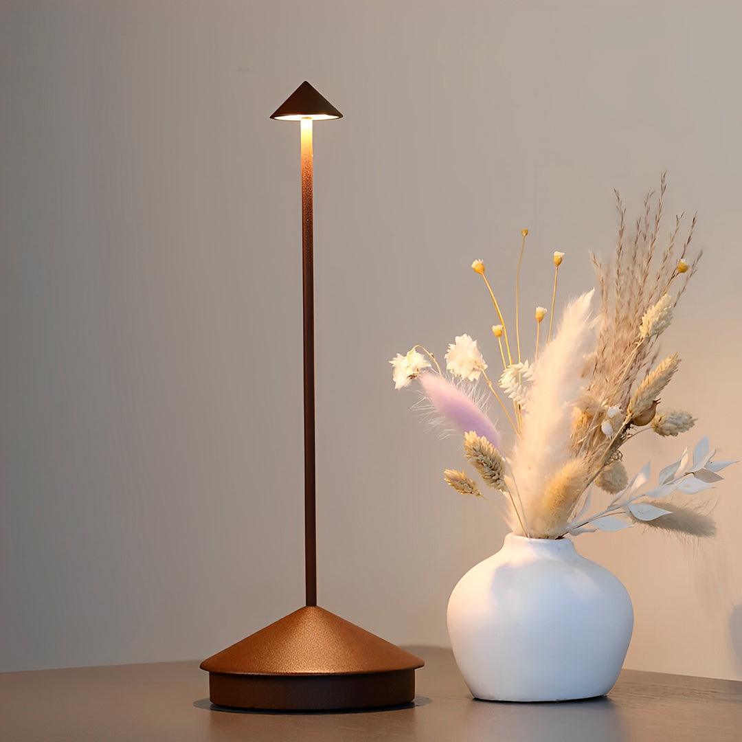Luminária marrom acesa ao lado de um vaso branco com flores secas, em fundo claro