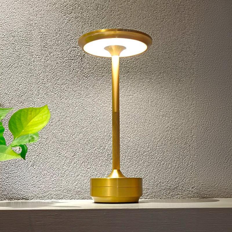 Luminária de mesa touch com disco de metal acesa sobre balcão, ao lado de folhas