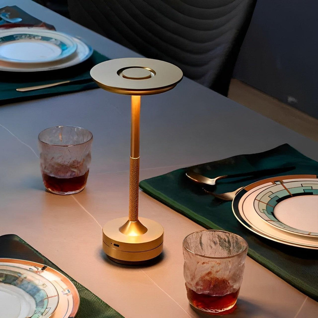 Peça dourada acesa sobre uma mesa posta, entre copos, pratos e jogo americano verde